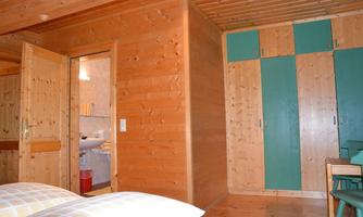 A cozy room with wooden walls and a large wardrobe. In the background, a bathroom and a simple table can be seen.