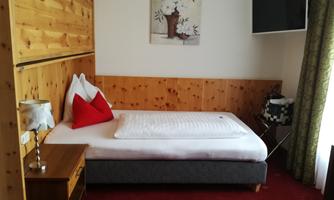 A cozy single room with wooden paneling and a modern television. On the bed are decorative cushions and a nightstand with a lamp stands beside it.