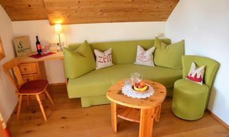 A cozy living room with a green couch and a round wooden table. On the table, there is fruit and a bottle of wine, accompanied by a warm wooden blanket and an armchair.