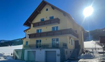 A yellow house with several balconies and garages. In the background, a snow-covered landscape and a clear blue sky can be seen.
