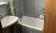 A small bathroom with a bathtub, a sink, and gray tiles. The door is open and provides a view of the room.