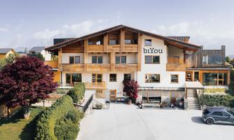 A modern three-story wooden building with the inscription "biYou". In front of the house, there are green areas and a parking area.