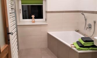 A modern bathroom with a bathtub and green towels. Bright walls and a large window opening create a friendly atmosphere.
