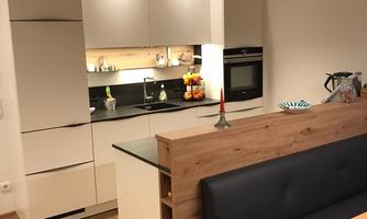 A modern kitchen with white cabinets and a wooden countertop. In the foreground, there is a cozy dining area with a black sofa and a wooden table.