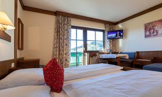 A cozy bedroom with two beds and a red decorative pillow. Large windows provide a beautiful view outside and a TV corner is also available.