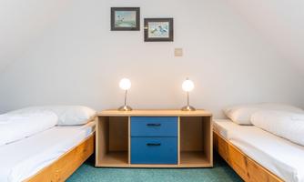 A cozy room with two single beds and a wooden piece of furniture in the middle. Two table lamps provide soft lighting and two pictures hang on the wall.