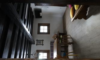 A cozy room with a wooden ceiling and windows. A dining table with chairs and colorful cushions on a bench are visible.