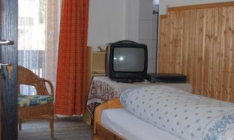 A simple room with a bed, a chair, and a television. The walls are light-colored and there are orange curtains.