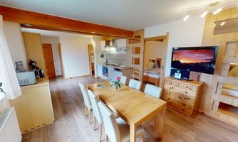 A modern living room with a wooden table and chairs. In the background, there is a kitchenette and a television visible.