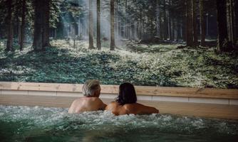 A couple relaxes in the water while gazing at a wall with a forest motif. The atmosphere is calm and inviting.