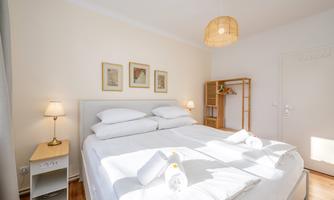A bright bedroom with a large bed, two pillows, and a cozy atmosphere. On the wall hang some pictures, and there is a bright rug as well as a small wardrobe.