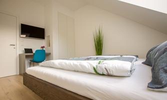 A modern bedroom with a cozy bed and a desk. The walls are light, and the furnishings are simple and functional.