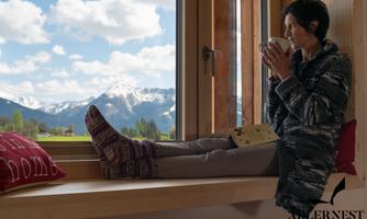 A woman is sitting comfortably by the window, enjoying a hot drink. In the background, there are snow-covered mountains and a blue sky with clouds.