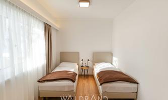 A bright room with two single beds and a warm wooden floor. Large windows let in plenty of daylight, giving the room a friendly atmosphere.