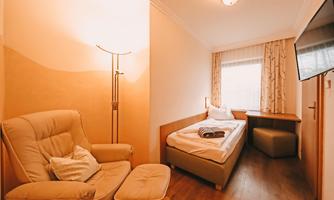 A cozy hotel room with a single bed and an armchair. The room is brightly lit with a window and a small table.