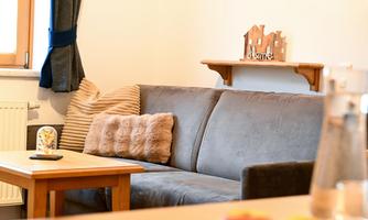 A cozy living room setup with a gray sofa and a wooden table. In the background, decorative elements and windows with curtains are visible.