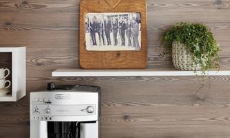 A modern coffee machine stands on a stylish kitchen countertop. In the background, there are some shelves with glasses and a photograph, as well as a basket with croissants and a plant.