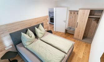A modern bedroom with a large bed. The room has light wooden paneling and a wardrobe.