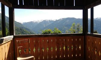 A view of the mountains with snow-capped peaks and plenty of greenery. In the foreground, there is a wooden chair in a covered area.