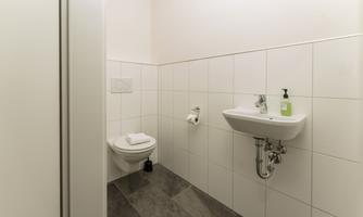 A modern bathroom with white tiles and a sink. There is a toilet and a green soap on the sink.