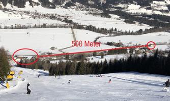 A wintry view of a ski slope that extends over 500 meters. In the background, snow-covered hills and a small village can be seen.