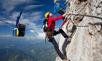 Two climbers are ascending a steep rock face. In the background, a cable car can be seen traveling through a picturesque mountain landscape.
