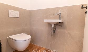 A modern bathroom with a wall-mounted toilet and a small sink. The walls are designed with elegant gray tiles and a wooden floor.