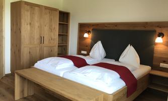 A cozy bedroom with a wooden bed and white pillows. In the background, there is a large wooden wardrobe and soft lighting.