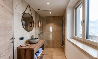 A modern bathroom with a shower and a round mirror. The decor is made of wood and stone, with plenty of daylight coming through the window.