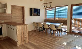 A modern living room with wooden decor and large windows. A dining table with chairs and an open kitchen area are visible.