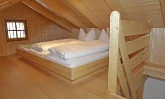 A cozy attic room with a wooden bed and two white pillows. The walls and floors are made of light wood, and a small window provides daylight.