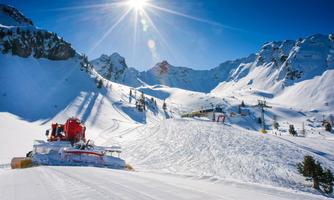 A snow-covered mountain landscape with a groomed slope section. In the background, ski lifts and tall mountains can be seen under a bright blue sky.