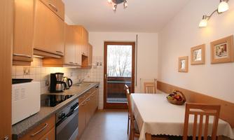 A modern kitchen with wooden cabinets and a bright countertop. A dining table with chairs and a fruit bowl is in the foreground.