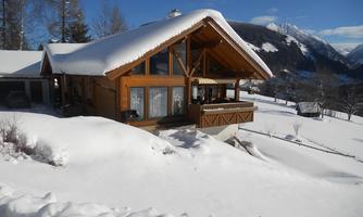 A beautiful wooden house in the snow, surrounded by mountains and trees. The sky is clear and blue.