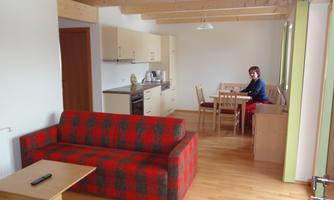 A modern living room with a red sofa and a bright kitchen. At the table sits a person working.