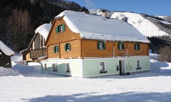 A charming wooden house with green shutters in a snowy landscape. Snow-covered mountains can be seen in the background.