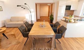 A modern living room with a large dining table and six chairs. There is also a cozy sofa and an open kitchen in the background.