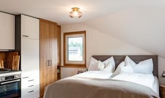 A modern bedroom with a large bed and wooden floor. The walls are light and there is a kitchenette in the background.