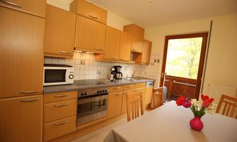 A modern kitchen with wooden furniture and a dining area. Large windows provide a view outside.