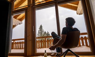 A man sits relaxed in a modern chair and looks through large windows at a winter landscape with snow-covered trees. Next to him, a bottle of wine stands on the floor.