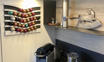 A modern kitchen with a Nespresso machine and a milk frother. Coffee capsules hang on the wall and decorative figures are placed on the countertop.