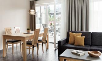 A modern living room with a cozy sofa and a dining table. Large windows provide plenty of natural light and a view of the terrace.