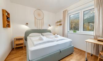 A cozy bedroom with a large bed and wooden flooring. The room has windows with a view and a decorative wall design.
