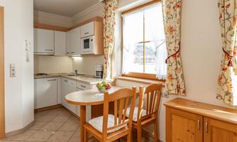 A bright kitchen corner with modern furniture and large windows. There is a small dining table with chairs and a beautifully decorated wooden cabinet.