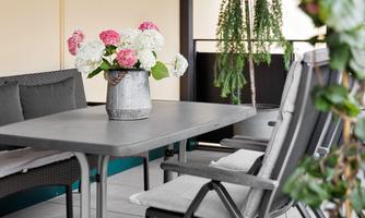 A cozy balcony with a gray table and chairs. On the table sits a vase with colorful flowers and there are some plants in the background.