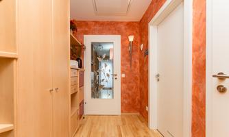 A bright hallway with orange walls and wooden floor. On the left side, there are cabinets and a door leads to another room.