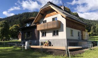 A modern house with a balcony and a large terrace.  
Surrounded by trees and mountains, in nice weather.