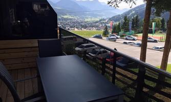 A balcony with a table and chairs that offers a beautiful view of the mountains and the valley. In the background, trees and parked cars can be seen.