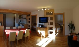 A modern living room with a dining table and six chairs. The open kitchen is well-lit, and a television is mounted on the wall.
