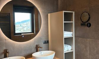 A modern bathroom with double sinks and a large round mirror. In the background, towels and a window with a view can be seen.